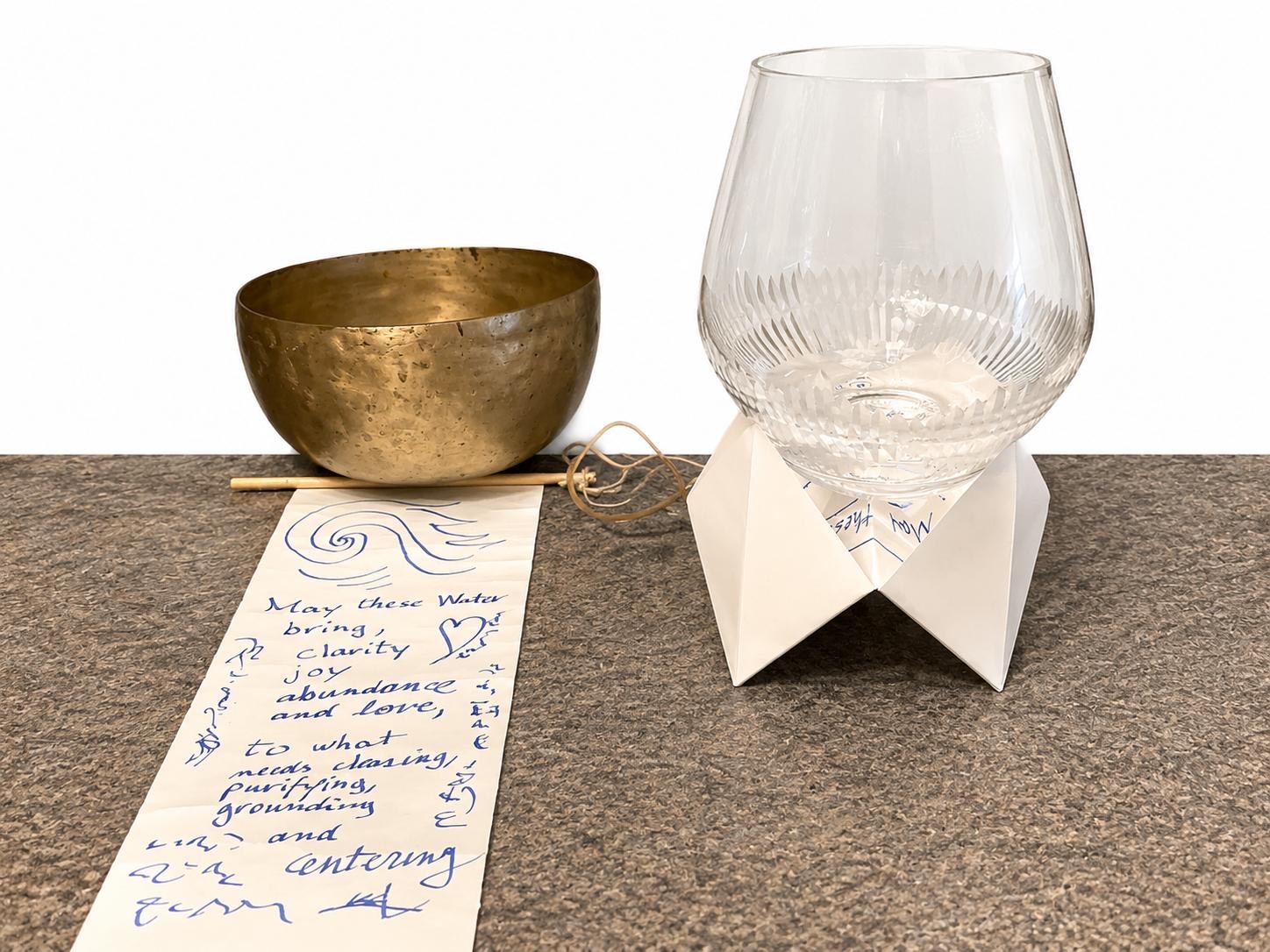The apparatus assembled: a hammered brass singing bowl with its wooden mallet, a cut-glass vessel of water resting inside a white folded-paper stand, and a paper scroll unrolled across the bench covered in blue handwriting and a drawn wave.