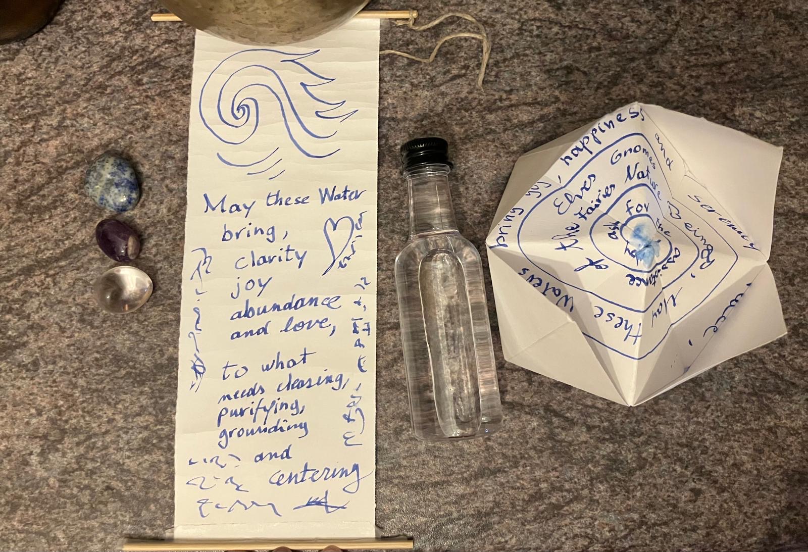 The components laid out flat on a granite bench for inspection: three tumbled stones — blue sodalite, purple amethyst, and clear quartz — beside the unrolled scroll with its drawn wave and blue handwriting, a small bottle of water, and the folded paper vessel covered in inked words spiralling toward its centre.